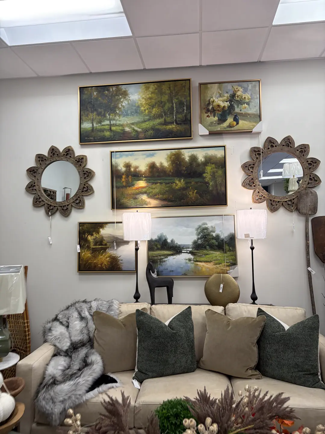 A sofa with green and taupe pillows under a wall of landscape paintings and mirrors